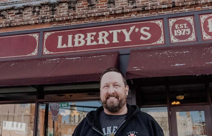 Smiling man stands outside Libertys Bar in Minnesota, next to a sign reading Est. 1975.