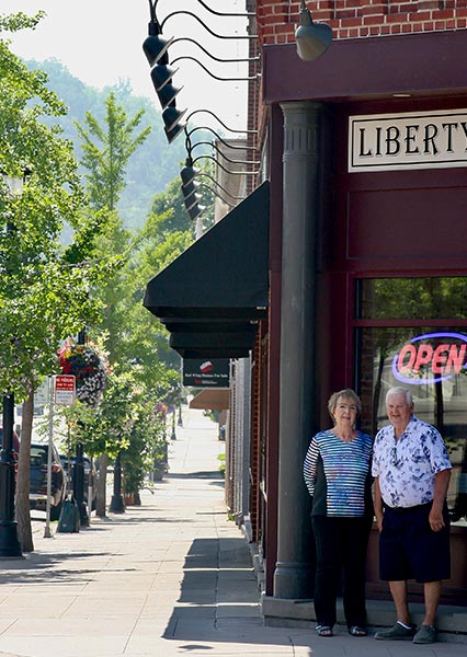 Two elderly people stand outside a Minnesota restaurant with an open sign on a sunny sidewalk.